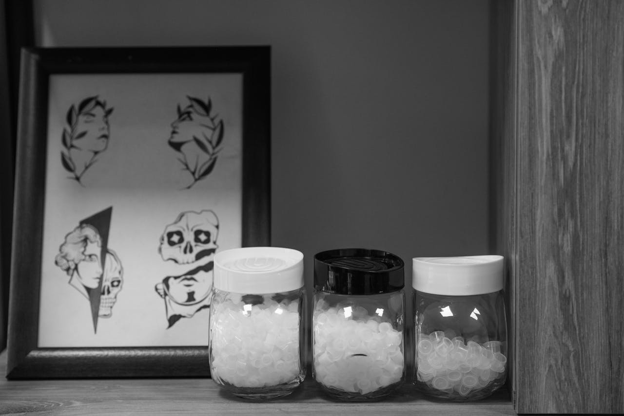 Black and white image of tattoo supplies including jars and framed art in a studio setting.